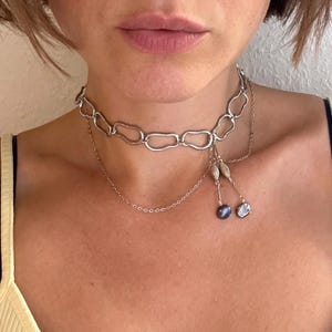 Op de afbeelding: Een zilverkleurige ketting met een chunky kettingontwerp. De ketting heeft een fijne ketting met hangende accenten, waaronder kleine kralen en twee iriserende, druppelvormige hangers. De sieraden worden om de nek gedragen.