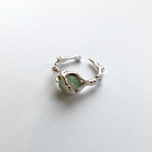 Pu&ograve; includere: Un anello color argento con una pietra preziosa verde chiaro. L'anello ha un design unico e organico, con una fascia che sembra fatta di piccoli rami. La pietra preziosa rotonda &egrave; incastonata al centro.