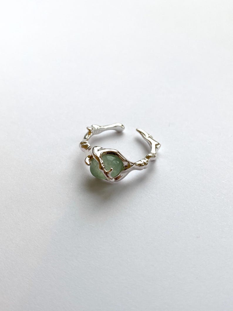 Pu&ograve; includere: Un anello color argento con una pietra preziosa verde chiaro. L'anello ha un design unico e organico, con una fascia che sembra fatta di piccoli rami. La pietra preziosa rotonda &egrave; incastonata al centro.