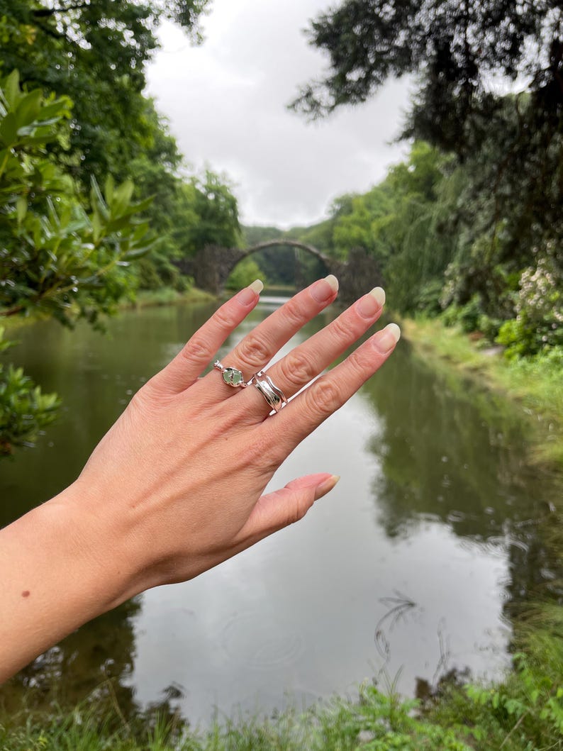 Pu&ograve; includere: Una mano che indossa due anelli d'argento, uno con una pietra verde. Gli anelli sono esposti sullo sfondo di un ponte di pietra sull'acqua, circondato da vegetazione lussureggiante. La scena &egrave; ambientata all'aperto in una giornata nuvolosa.