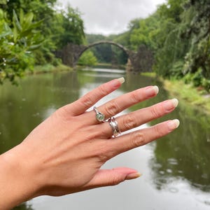 Pu&ograve; includere: Una mano mostra due anelli: un anello d'argento con una pietra preziosa verde chiaro e una fascia d'argento con un design ondulato. Lo sfondo presenta un ponte di pietra sull'acqua, circondato da alberi verdi.