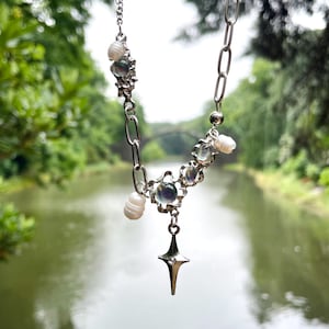 Könnte beinhalten: Eine silberfarbene Halskette mit einem Sternanhänger und einem Kettenglied-Design. Die Halskette hat schillernde florale Akzente und kleine, weiße Perlen. Der Hintergrund zeigt einen Fluss und Bäume.