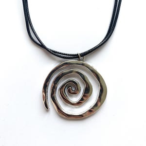 Könnte beinhalten: Halskette mit silberfarbenem Anhänger in Spiralform. Der Anhänger hängt an einer schwarzen Kordelkette. Das Spiraldesign hat eine gehämmerten Textur, vor weißem Hintergrund.