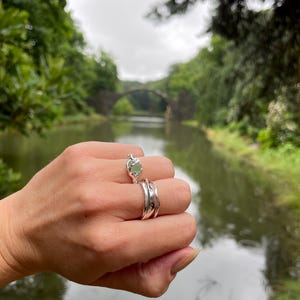 Pu&ograve; includere: Una mano indossa tre anelli d'argento. L'anello superiore presenta una pietra preziosa verde chiaro. Gli altri due anelli hanno un design ondulato. Sullo sfondo, un ponte sull'acqua.