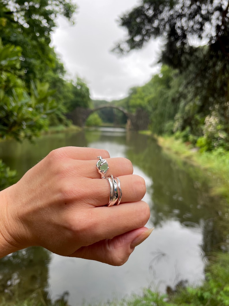 Pu&ograve; includere: Una mano indossa tre anelli d'argento. L'anello superiore presenta una pietra preziosa verde chiaro. Gli altri due anelli hanno un design ondulato. Sullo sfondo, un ponte sull'acqua.