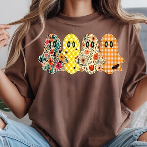 Peut inclure: Un t-shirt marron à manches courtes orné d'un motif représentant quatre fantômes alignés. Chaque fantôme présente un motif distinct : fleurs colorées, vichy jaune, imprimés botaniques floraux et vichy orange, avec des contours cousus.