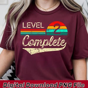 May include: A maroon short-sleeved t-shirt featuring a graphic print with the text "LEVEL 2 Complete". The design includes a distressed, multi-colored sunset arc in red, orange, and teal, with a large number two and a decorative underline.