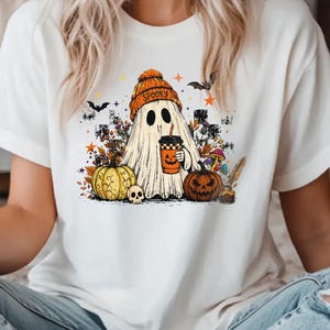 Peut inclure: Un t-shirt blanc avec un motif représentant un fantôme portant un bonnet orange sur lequel est écrit SPOOKY. Le fantôme tient une tasse de café orange, entouré d'une citrouille jaune, d'une citrouille d'Halloween, d'un crâne et de feuilles.