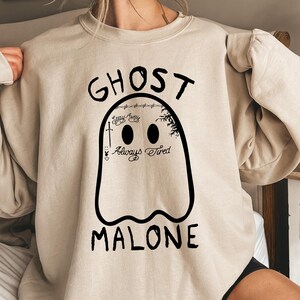 May include: A beige sweatshirt with a black ghost graphic. The ghost has two black eyes and the words "Ghost Malone" printed below. Above the ghost are the words "Stay Away" and "Always Tired".