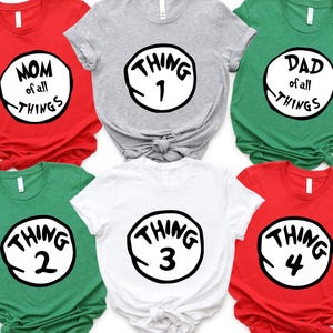 May include: Six short-sleeved t-shirts in red, gray, green, and white. Each shirt features a black and white circular design with the words "MOM of all THINGS", "THING 1", "DAD of all THINGS", "THING 2", "THING 3", and "THING 4".