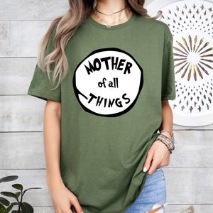 May include: Olive green t-shirt with the text "MOTHER of all THINGS" in a black and white circular design. The shirt is a crew neck style and is made of a soft material. The model is wearing blue jeans.