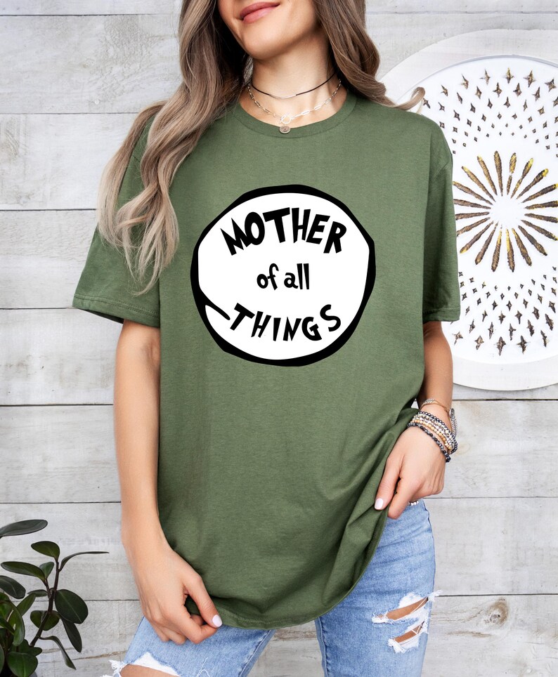 May include: Olive green t-shirt with the text "MOTHER of all THINGS" in a black and white circular design. The shirt is a crew neck style and is made of a soft material. The model is wearing blue jeans.