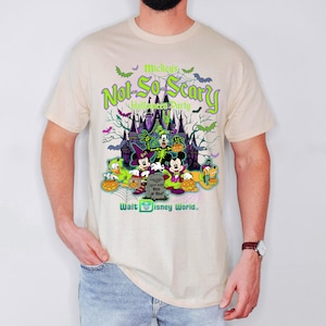May include: Beige t-shirt featuring a colorful graphic for Mickey's Not-So-Scary Halloween Party at Walt Disney World. The design includes Mickey Mouse, Minnie Mouse, Donald Duck, and Goofy in Halloween costumes, with a spooky castle backdrop.