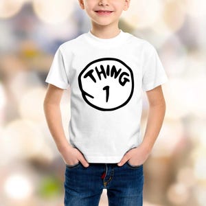 May include: White t-shirt with a black graphic that says "THING 1" inside a black circle. The shirt is worn by a child with hands in their pockets, wearing blue jeans. The background is blurred.