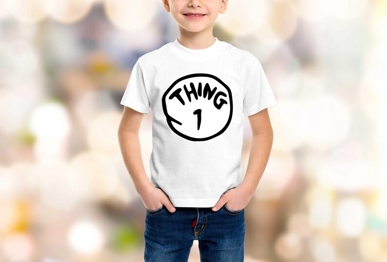 May include: White t-shirt with a black graphic that says "THING 1" inside a black circle. The shirt is worn by a child with hands in their pockets, wearing blue jeans. The background is blurred.