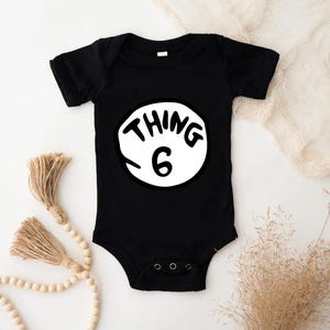 May include: Black baby bodysuit with the text "THING 6" in a white circle. The text is in a playful, handwritten style. The bodysuit has short sleeves and a snap closure.