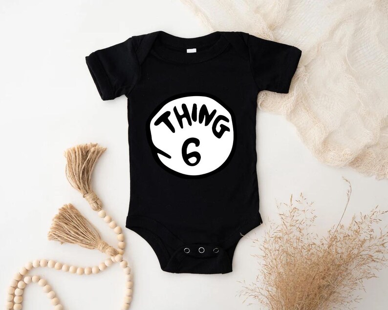 May include: Black baby bodysuit with the text "THING 6" in a white circle. The text is in a playful, handwritten style. The bodysuit has short sleeves and a snap closure.