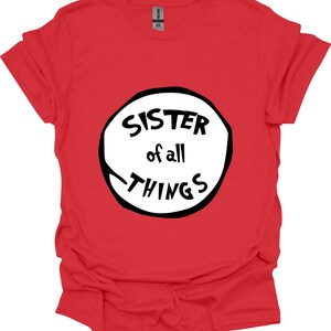 May include: A red t-shirt with the text "SISTER of all THINGS" in a black and white design. The text is inside a white circle with a black outline. The shirt is made of a soft material.