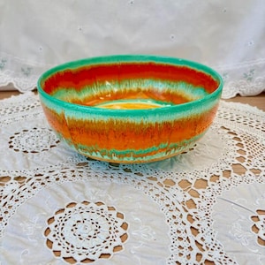 May include: A ceramic bowl with a vibrant color scheme. The bowl features a gradient of colors, including orange, yellow, and green. The bowl is placed on a white lace doily.