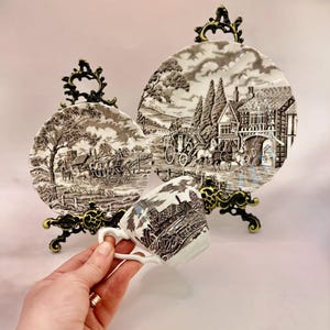 May include: A set of decorative plates and a teacup with a brown and white pastoral scene. The plates are displayed on ornate, dark metal stands. The teacup features the same scenic design.