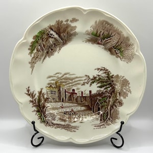 May include: A white ceramic dinner plate with a brown and green landscape design featuring a castle, trees, and a horse-drawn carriage. The plate has a scalloped edge.