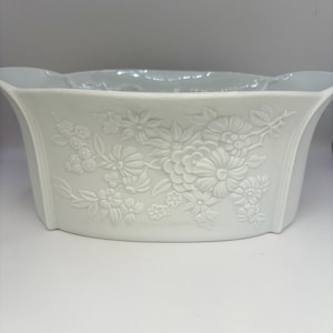 May include: A white ceramic planter with a floral relief pattern. The rectangular planter has a gently flared shape and a smooth, matte finish. The embossed flowers and leaves provide a touch of elegance to this decorative item.
