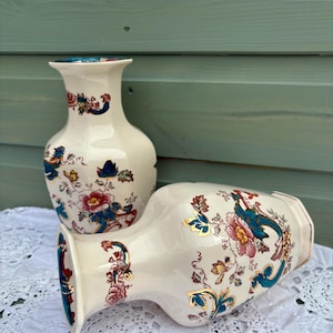 May include: Two cream-coloured ceramic vases. They feature floral patterns in red, blue, and gold. One vase stands upright, the other lies on its side. The vases have a classic shape and are decorated with detailed botanical designs.