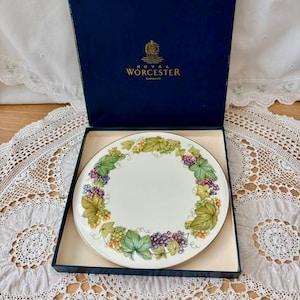 May include: A round, white Royal Worcester plate with a gold rim, featuring a grape and leaf design. The plate is in a navy blue box with the Royal Worcester logo. The plate is approximately 10 inches in diameter.