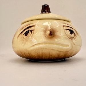 May include: A ceramic onion-shaped container with a lid, depicting a sad face. The container is beige with brown shading and accents, including a dark brown knob on the lid. The face has tear-shaped details. A decorative kitchen item.
