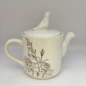 May include: Cream ceramic teapot with a bird-shaped lid handle. The teapot has a brown floral design. The spout and handle are curved. A charming addition to any kitchen or tea set. The teapot is a lovely item.