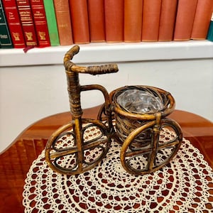 May include: A decorative tricycle-shaped basket made of woven brown material. The tricycle has three wheels and a basket in the front. It sits on a cream-coloured, circular doily. A shelf of antique books is in the background.