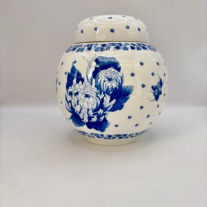 May include: A white ceramic ginger jar with a lid, adorned with blue floral designs. The jar features a large blue flower motif and small blue dots. The lid also has blue accents, creating a cohesive decorative piece.