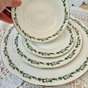 May include: A white porcelain dinner service featuring a green and gold leaf pattern. The set comprises a bowl, a side plate, and two dinner plates. The edges are finished with gold trim.