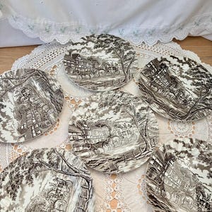 May include: Set of six vintage ceramic plates with a black and white transferware design. Each plate features a detailed landscape scene with buildings, trees, and horse-drawn carriages. The plates are arranged on a white lace doily.