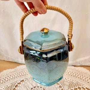 May include: A vintage ceramic jar with a light blue glaze and gold accents. The jar has a lid and a woven handle. The jar is decorated with a subtle pattern and is likely used for storage.