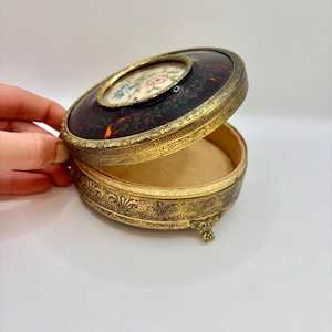 May include: An antique jewelry box with a hinged lid. The lid is dark brown with a tortoiseshell pattern and a gold-colored frame. The top features a small oval image of flowers. The base is gold-colored with ornate detailing.