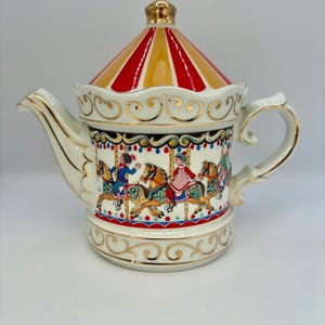 May include: A white porcelain teapot with a carousel design. The teapot has a gold trim and a red and yellow striped lid. The carousel features horses and riders in colorful clothing.