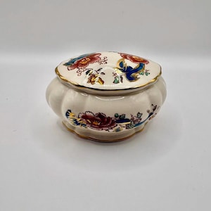 May include: A small, cream-colored ceramic trinket box with a lid. The lid and base are adorned with floral patterns in red, blue, and gold. The box has a gold trim and a rounded shape, suggesting a vintage or antique style.