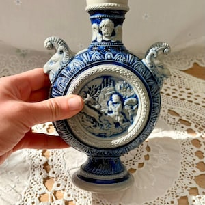 May include: A blue and white ceramic flask with a detailed relief scene. The flask features ram's head handles and a decorative rim. The central relief depicts figures in a landscape. The flask is approximately 8 inches tall.