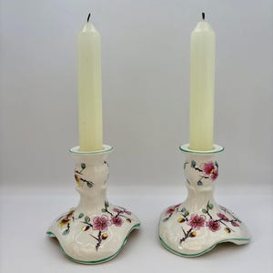 May include: Two cream-coloured ceramic candle holders with a floral design. Each holder has a flared base, a central stem, and a top ring, with a white candle in each. The floral design includes pink blossoms and green leaves.