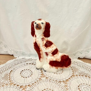 May include: A ceramic dog figurine in a seated position. The dog is mainly white with reddish-brown markings on its ears, head, and body. The figurine has a detailed, hand-painted appearance, with a glossy finish. The dog is sitting on a white lace doily.