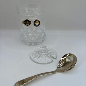 May include: A clear crystal jar with a diamond pattern, a lid, and a silver spoon. The jar has a label that reads "Royal Brierley Crystal". The spoon is curved with a rounded bowl.