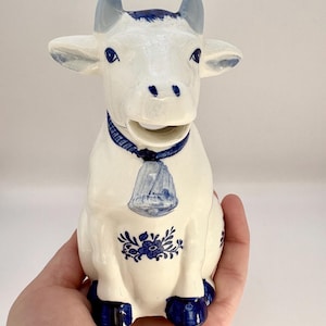 Könnte beinhalten: Ein weißer Keramik-Milchkännchen in Form einer Kuh mit blauen und weißen Delftware-Designs. Die Kuh hat eine blaue Glocke um den Hals und blaue Hufe.