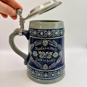 May include: A blue ceramic beer stein with a hinged metal lid. The stein features a decorative design with the words "Hopfen u. Malz Gotterhalt's" in white. The handle and lid are silver-coloured. The stein is approximately 18 cm tall.