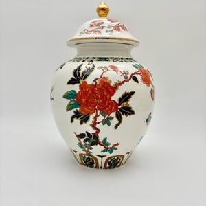 May include: A white ceramic jar with a lid, featuring a floral design in red, orange, and green. The jar has a gold finial and gold trim. The floral pattern includes a large red flower and branches.