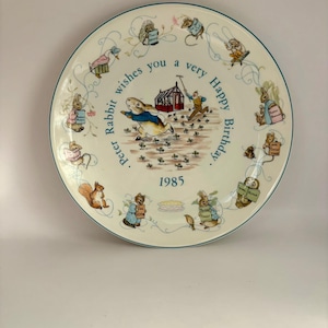May include: A vintage ceramic plate with a teal rim, featuring illustrations of Peter Rabbit and other animals. The plate has the text "Peter Rabbit wishes you a very Happy Birthday 1985" around the central image.