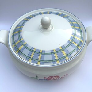 May include: A white ceramic casserole dish with a lid featuring a blue, yellow, and green plaid pattern. The dish has two handles and a small floral design on the side. The lid has a white knob.