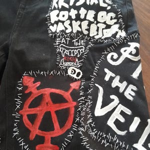 May include: Black denim shorts with white and red painted patches. Patches include text that reads "KRYSTAL ROTTE OG VASKERIJ", "EAT THE RICH", and "F THE VEIL". The shorts have a punk rock aesthetic.