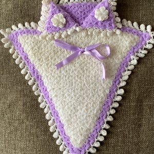 Può includere: Un cuscino bianco all'uncinetto con un bordo all'uncinetto viola e un nastro di raso viola. Il cuscino ha una forma a stella ed è decorato con fiori bianchi all'uncinetto.