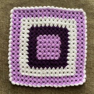 Può includere: Un tovaglietta all'uncinetto quadrata con un motivo viola e bianco. La tovaglietta è realizzata in filato e ha un design delicato e traforato.